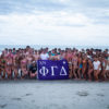 Large group of young people in swimsuits posing on a beach, holding a purple banner with Greek letters (XN ΦΓΔ).