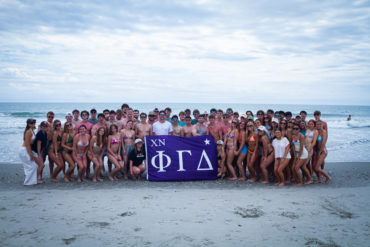 Large group of young people in swimsuits posing on a beach, holding a purple banner with Greek letters (XN ΦΓΔ).