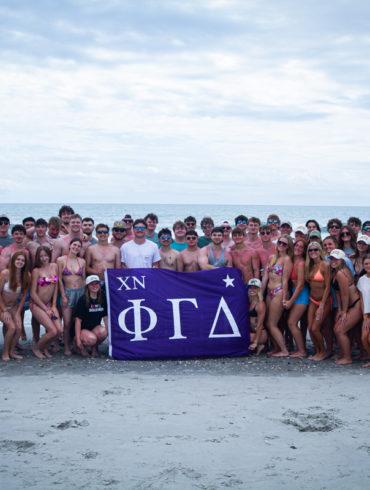 Large group of young people in swimsuits posing on a beach, holding a purple banner with Greek letters (XN ΦΓΔ).