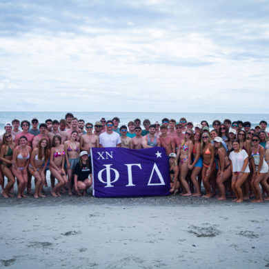 Large group of young people in swimsuits posing on a beach, holding a purple banner with Greek letters (XN ΦΓΔ).