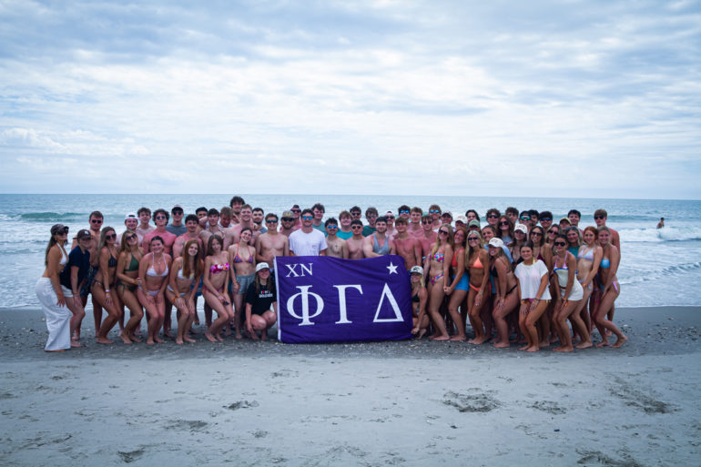 Large group of young people in swimsuits posing on a beach, holding a purple banner with Greek letters (XN ΦΓΔ).