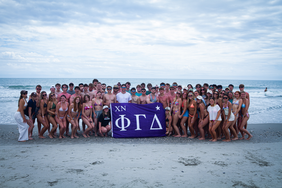 Large group of young people in swimsuits posing on a beach, holding a purple banner with Greek letters (XN ΦΓΔ).