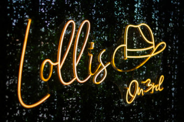 IMG_9782 | Beachmonkey Orange neon cursive sign glowing against a dark leafy background, close-up.